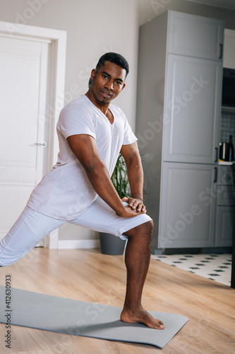 Serious fit African-American man making sport fitness exercise standing on yoga mat at bright domestic room, looking at camera. Concept of sport training at home gym.