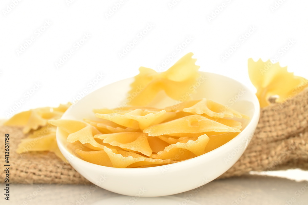 Light yellow uncooked pasta, in the shape of a butterfly, in a white dish with a jute bag, close-up, isolated on white. Light yellow uncooked pasta, in the shape of a butterfly, in a white dish with a jute bag, close-up, isolated on white.