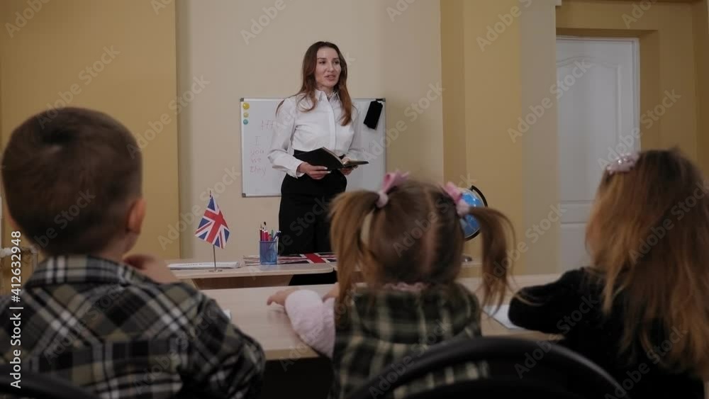Close-up of small children sitting at a desk in school and raising their hand to answer, the teacher smiles standing at the blackboard. Close-up of small children sitting at a desk in school and raising their hand to answer, the teacher smiles standing at the blackboard.