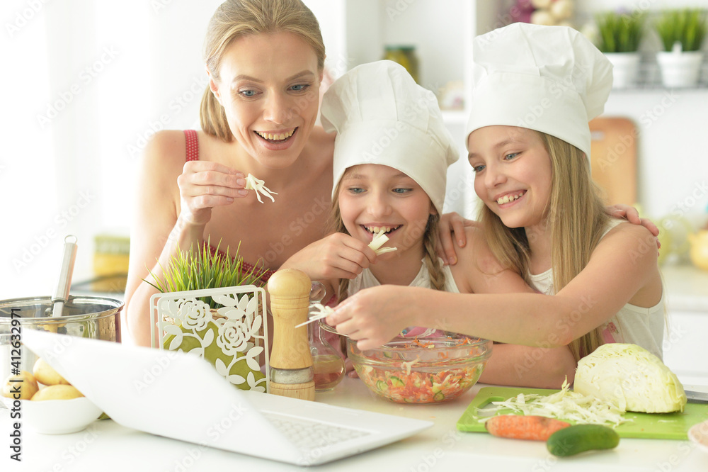 Cute girls with mother preparing delicious fresh salad in kitchen looking at laptop Cute girls with mother preparing delicious fresh salad in kitchen looking at laptop