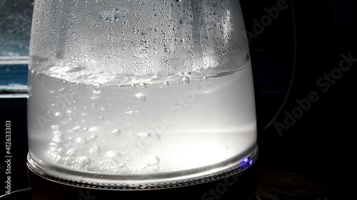 Defocused bubbles in hot water boiling inside glass teapot in bright sunlight. Closeup of boiling water in old transparent electric kettle on kitchen. Splashing water bubble background