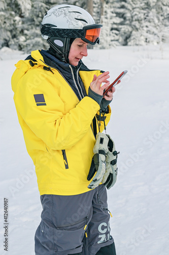 Woman snowboarder in bright suit in a sports helmet writes a message to smartphone. Cellular for extreme sports. Vertical shot