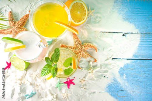 Summer tropical sea holiday vacation concept. Three various glass with summer cold cocktail drinks, on sand with seashells and starfish, light blue wooden background