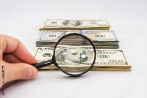 hand with a magnifying glass checks one hundred dollar bills against a white