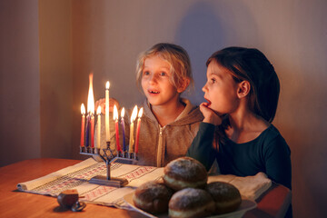 Girls friends lighting candles on menorah for traditional Jewish Hanukkah holiday at home. Children celebrating Chanukah festival of lights together. Dreidel and Sufganiyot donuts in plate on table.