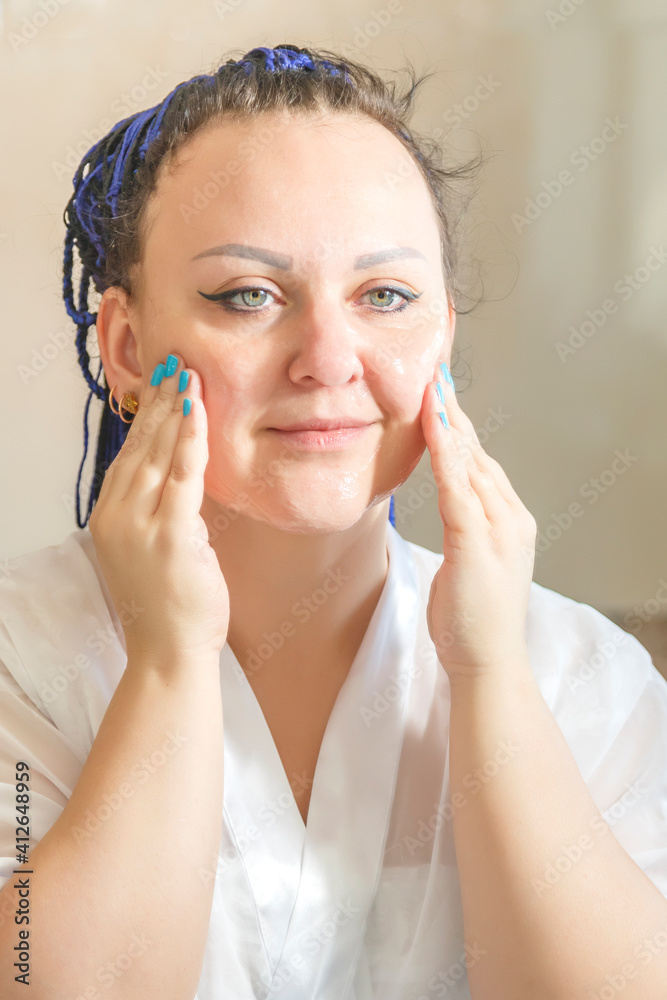 a woman in a white robe with a blue afro hairstyle in the bathroom near the mirror makes a face scrub. a woman in a white robe with a blue afro hairstyle in the bathroom near the mirror makes a face scrub.