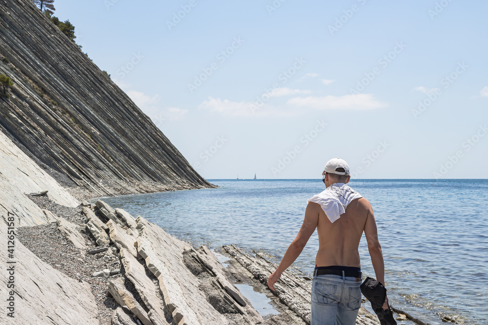 Beautiful summer seascape. A guy walks along a large stone wild beach. On the horizon, clear blue sky and steep cliffs. Surroundings of the resort city of Gelendzhik. Russia, Black Sea coast