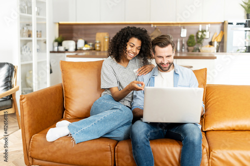 Fotografiet Cheerful diverse couple in love spends leisure time online with a laptop at home