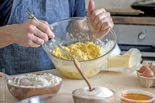 A faceless man in an apron kneads cookie dough with honey, flour and butter on the kitchen table with ingredients. Authentic hobby home baker. Close-up of a glass cup with dough and hands with a whisk