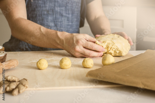 A man without a face in an apron makes homemade shortbread dough cookies on the kitchen table and rolls balls. Around ingredients for baking. Concept of authentic home hobby baker.