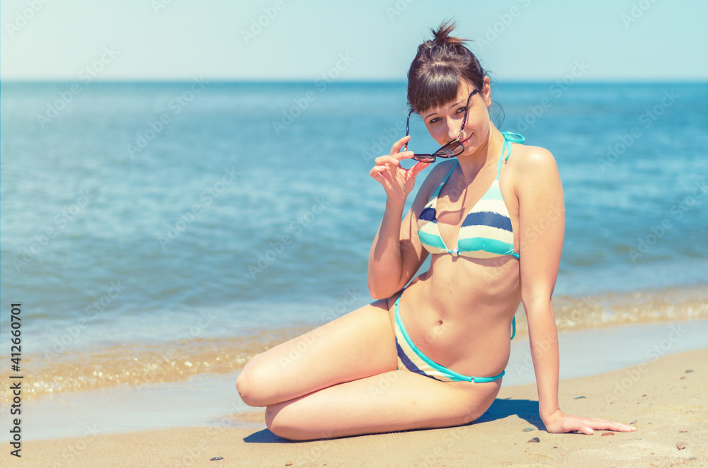 Young beautiful girl in bikini sit on sandy beach, put on sunglasses and looking at camera, blue water of Baltic sea, tourist female on vacation in Lithuania Curonian Spit in sunny summer day Young beautiful girl in bikini sit on sandy beach, put on sunglasses and looking at camera, blue water of Baltic sea, tourist female on vacation in Lithuania Curonian Spit in sunny summer day
