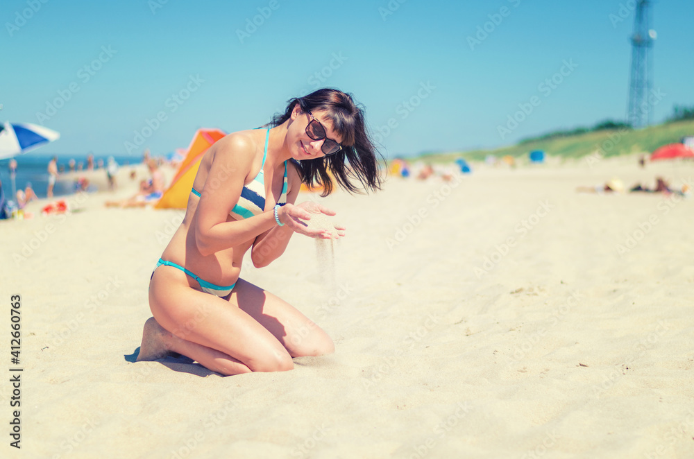 Young beautiful girl in bikini and sunglasses sit on sandy beach, pour sand through fingers and smile, Baltic sea water, tourist female on vacation in Lithuania Curonian Spit in sunny summer day