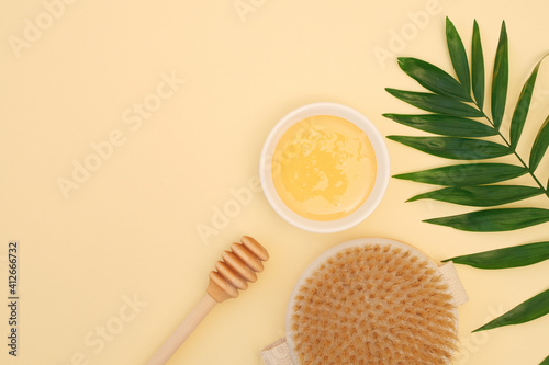 Honey in a bowl, honey dipper, dry massage brush, green leaf on a soft yellow background with space for text. Composition of ingredients for spa body skin care. Body Scrub. Anticellulite massage. Skin