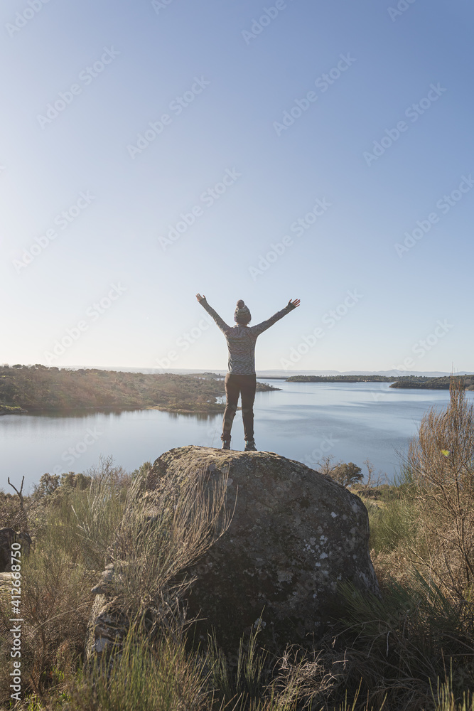 Young and attractive female with her arms opened and located on top of a rock with a lake in front of her during a sunny and cold winter morning. Young and attractive female with her arms opened and located on top of a rock with a lake in front of her during a sunny and cold winter morning.