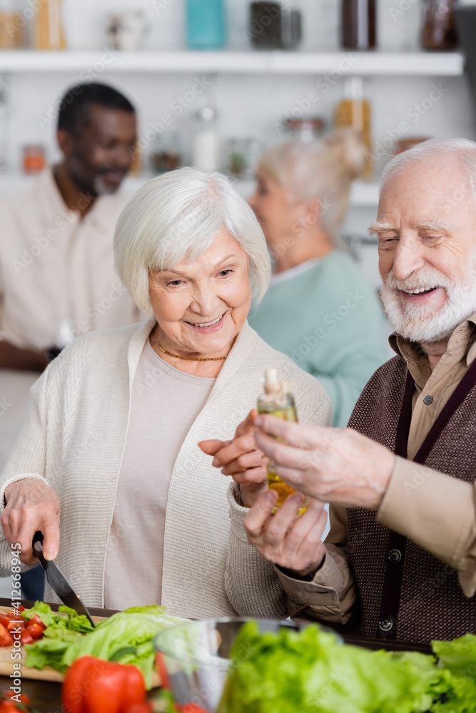 happy senior woman pointing with finger at bottle with oil near retired man and multicultural friends on blurred background happy senior woman pointing with finger at bottle with oil near retired man and multicultural friends on blurred background