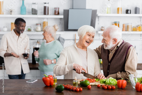 happy senior man and woman cooking salad near multiethnic friends on blurred background