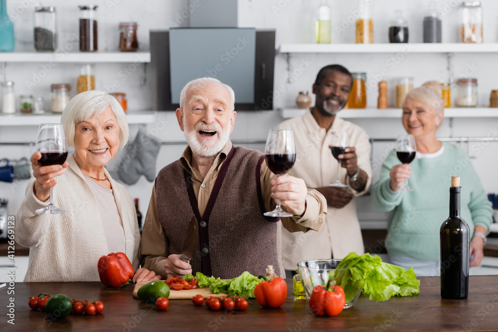 amazed senior man and woman holding glasses of wine near multicultural friends on blurred background amazed senior man and woman holding glasses of wine near multicultural friends on blurred background