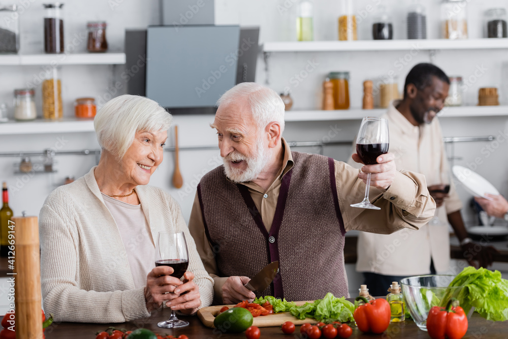 happy senior man and woman holding glasses of wine near multicultural friends on blurred background happy senior man and woman holding glasses of wine near multicultural friends on blurred background