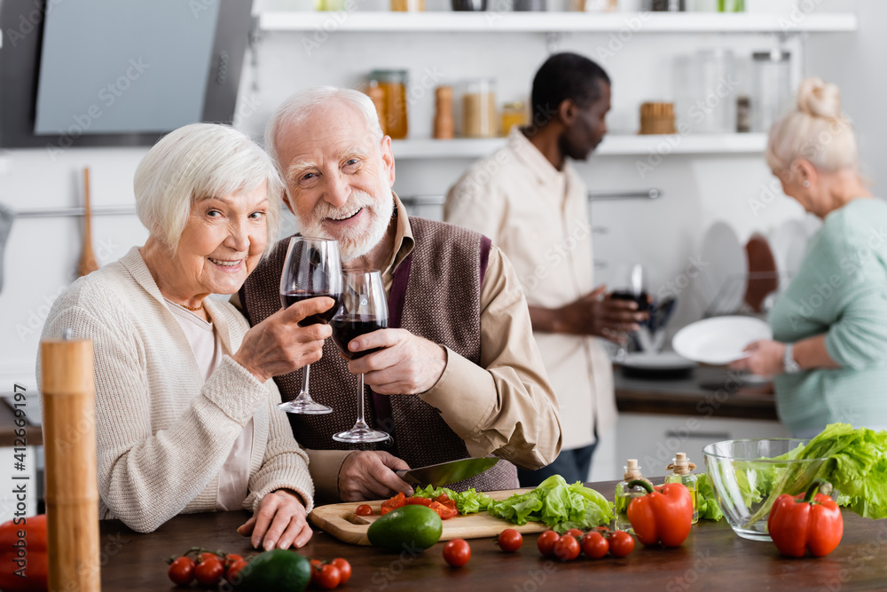 happy senior man and woman clinking glasses of wine near multicultural friends on blurred background