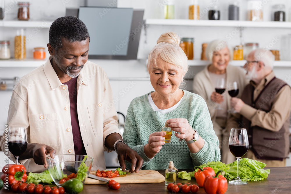 African american man and happy senior woman preparing salad near retired friends on blurred background