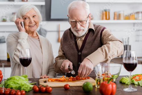 senior man cutting vegetables near happy retired wife talking on smartphone