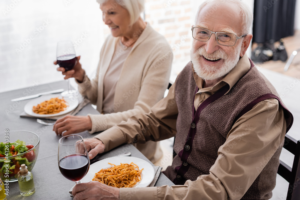 happy senior man in eyeglasses smiling while holding glass of wine during dinner with woman