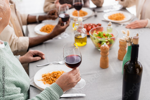 cropped view of senior woman holding glass of red wine near plate with pasta during lunch with multicultural friends