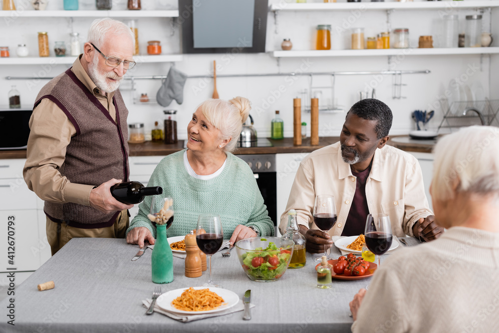 retired man in eyeglasses pouring red wine in glass near happy interracial friends retired man in eyeglasses pouring red wine in glass near happy interracial friends