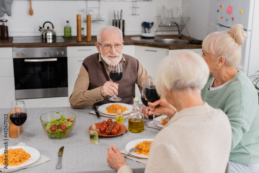 happy senior man clinking glasses of red wine with retired women near tasty lunch on table happy senior man clinking glasses of red wine with retired women near tasty lunch on table