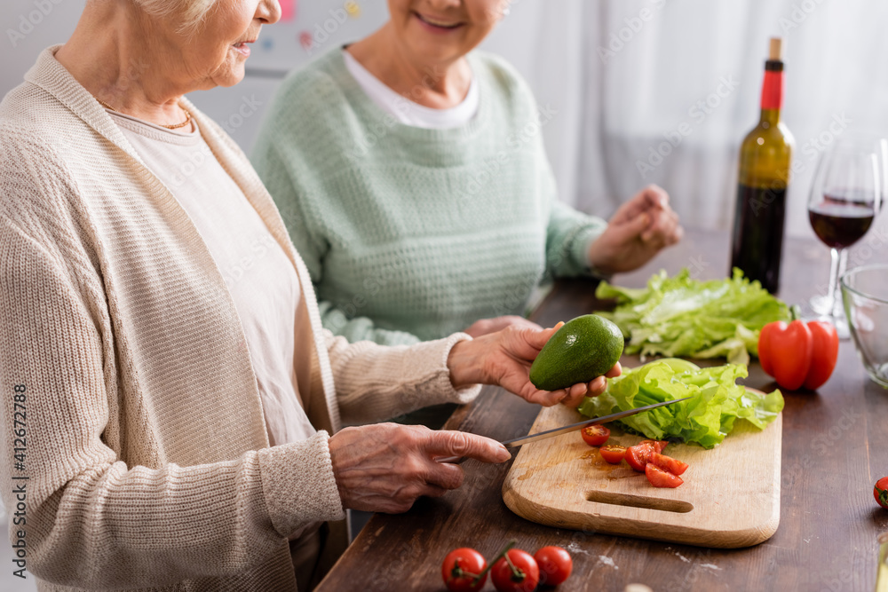 cropped view of senior woman holding knife and avocado near retired friend in kitchen