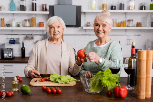 happy senior woman holding fresh bell pepper near retired friend in kitchen