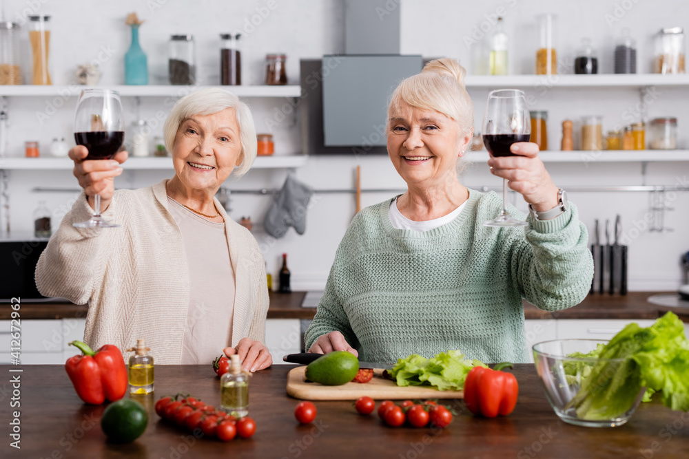 happy senior women holding glasses of red wine near fresh vegetables in kitchen happy senior women holding glasses of red wine near fresh vegetables in kitchen