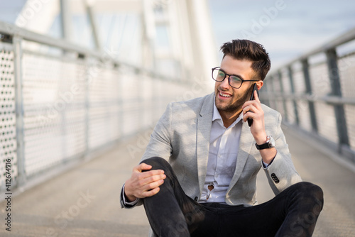 Happy young businessperson talking on smartphone sitting and relaxing on the bridge. Joyful yuppie or young entrepreneur outside photo