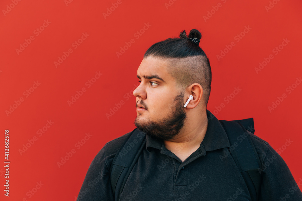 Fototapeta premium Bearded overweight man in wireless headphones looking to the side, close portrait on red background.
