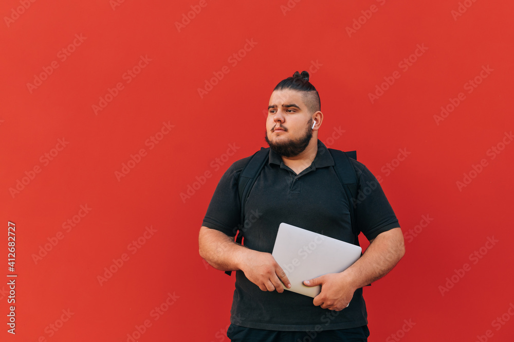 Serious man with a laptop in his hands and a backpack stands on the street against the backdrop of a red wall and looks to the side. Serious man with a laptop in his hands and a backpack stands on the street against the backdrop of a red wall and looks to the side.