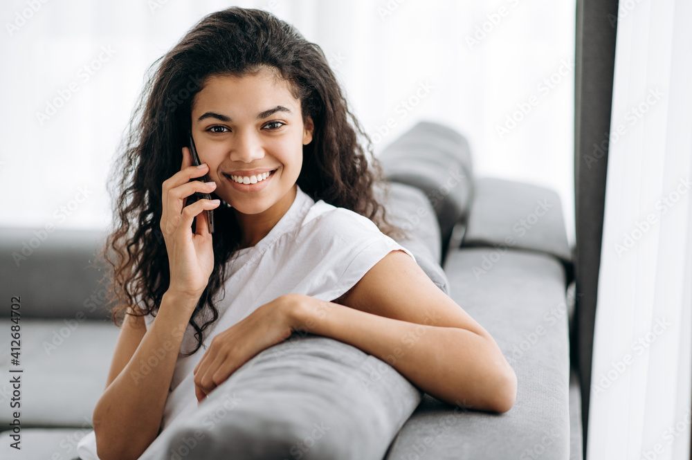 Smiling multiracial black curly girl uses cell phone, talking with friends or family while sits on the couch at living room and looks at camera, communication concept Smiling multiracial black curly girl uses cell phone, talking with friends or family while sits on the couch at living room and looks at camera, communication concept