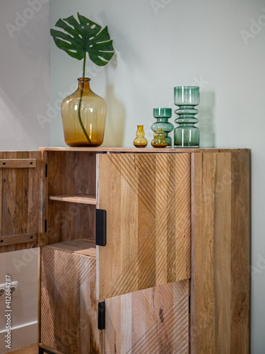 Transparent colorful vases for flowers and big bottle with monstera leaf on wooden cupboard with opened doors. photo