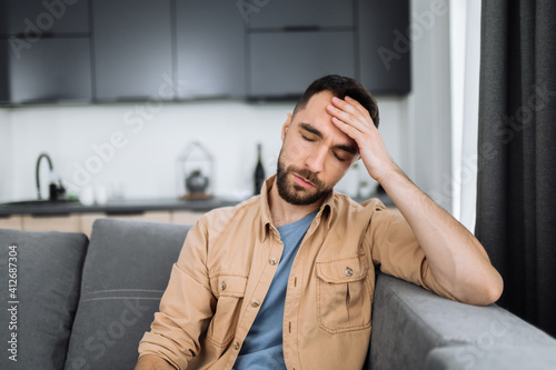 Tired caucasian bearded man sits on the sofa, having a headache. Unhealthy guy catch a cold or having flu, upset about illness, unhappy man feel depressed
