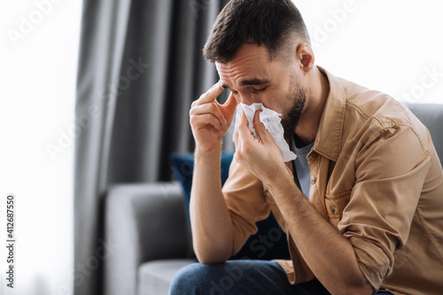 Unhealthy young man. Caucasian guy is sick with flu, he sneezes and blows his nose in a napkin while sitting on the couch at home