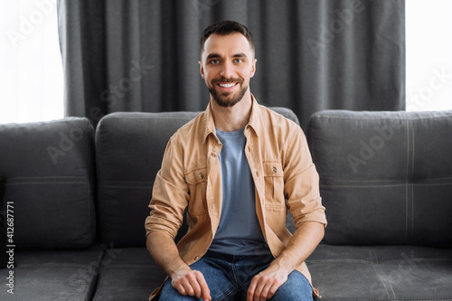 Portrait of an attractive caucasian bearded guy dressed in stylish casual wear. Confident man looks directly at camera with a friendly smile while sitting at the couch in a living room