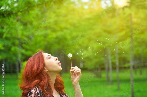 Pretty red-haired woman blows away a dandelion. Cute girl in a fashionable leopard print jacket. Romantic mood. Walk in the Park. Enjoy Nature. Green grass in summer Park.
