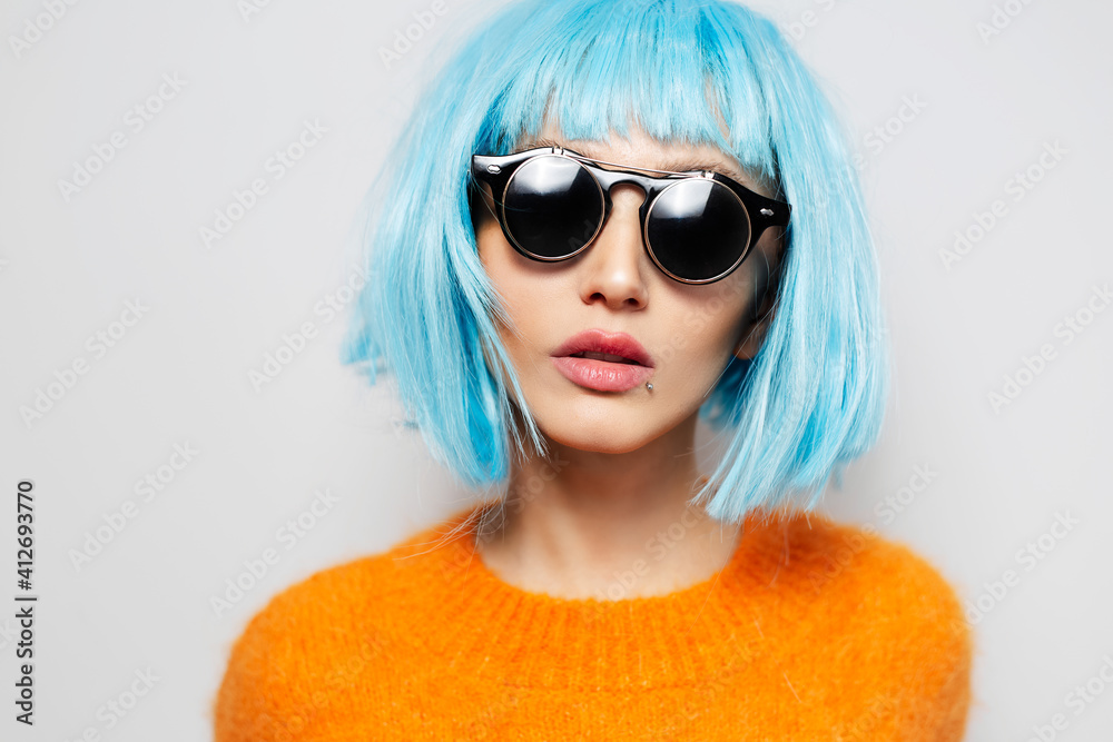 Studio close-up portrait of young girl with blue bob hairstyle, wearing sunglasses and orange sweater on white background. Studio close-up portrait of young girl with blue bob hairstyle, wearing sunglasses and orange sweater on white background.