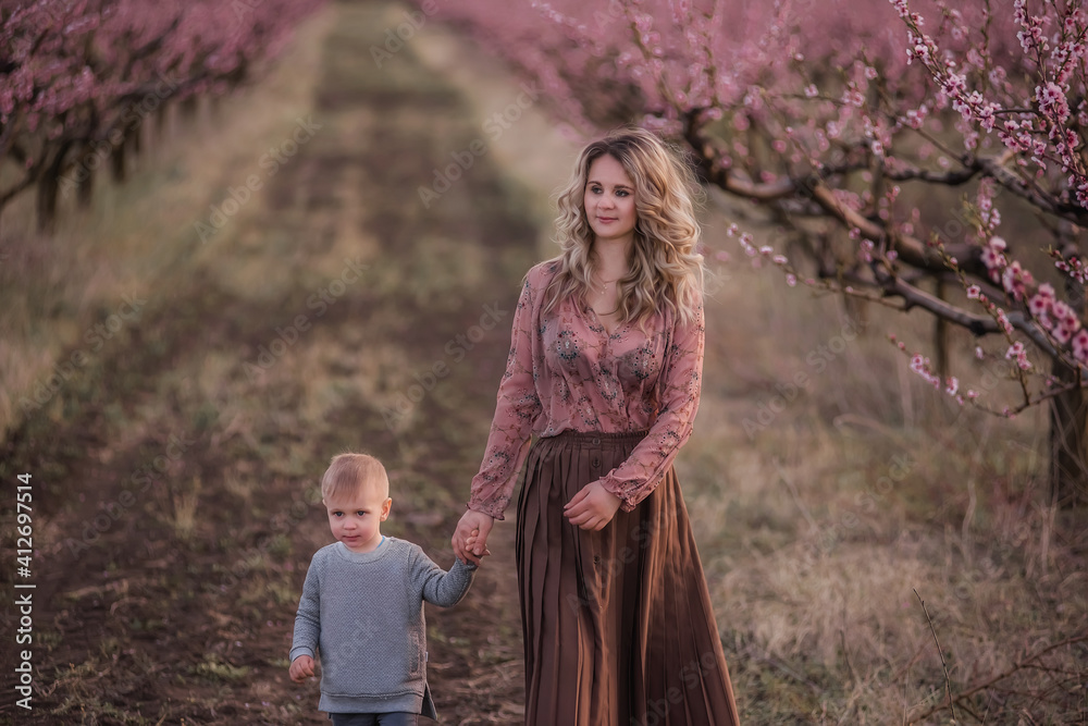 A young mother holds the hand of her little son, play, run together in the pink blossoming peach gardens. The woman hugs the boy, kisses him, waves her hand, calls him to go with her. Copy space