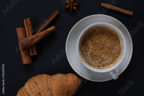 Cup of aromatic cappuccino with fresh croissant with cinnamon sticks on a dark background.