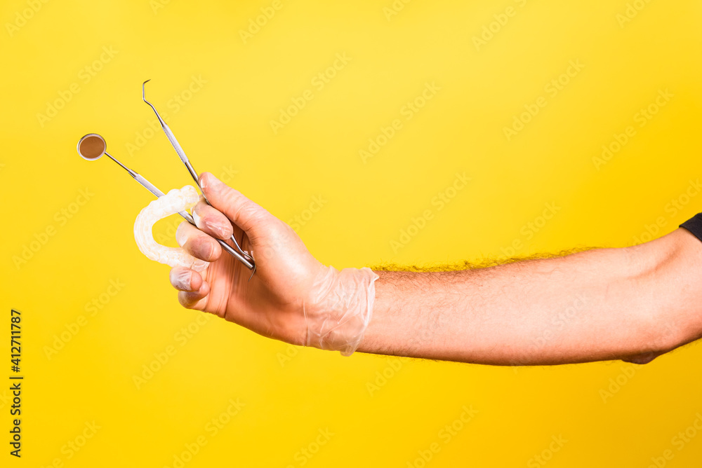 Dentist hand shows and holds mirror, scaler and a dental aligner, isolated on yellow background.