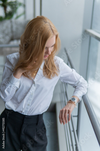 The girl is on the phone. with a kind look, on against the window, at the railing, in a white blouse looking at the watch