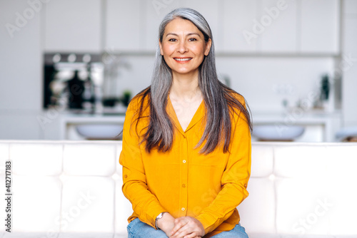 Portrait of a charming asian senior long gray hair lady wearing casual stylish clothes, sitting on couch at living room, looking directly at the camera with friendly smile