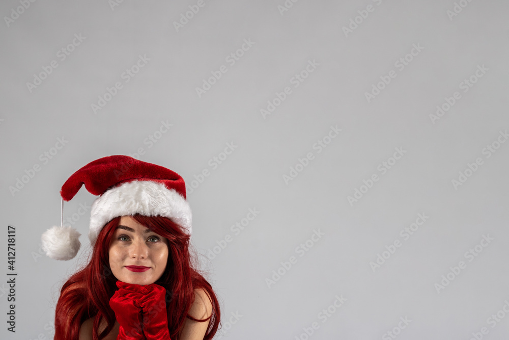 Young female attractive model with auburn hair and red lipstick sat down in a studio with a white background wearing a sexy santa outfit with a red Santa hat with red velvet gloves. Young female attractive model with auburn hair and red lipstick sat down in a studio with a white background wearing a sexy santa outfit with a red Santa hat with red velvet gloves.