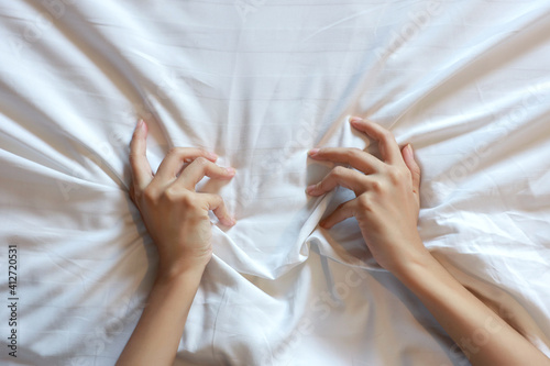 Top view young sexy woman hands pulling white sheets in ecstasy in hotel. Cute girl doing sign orgasm on white bed, sex and erotic concept for advertisement