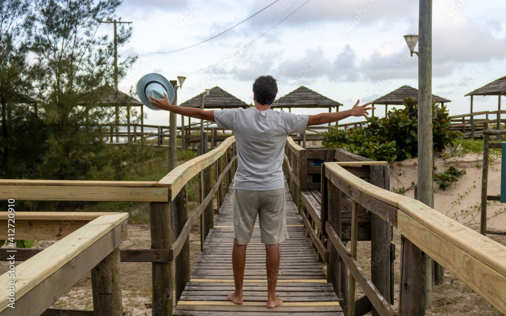 Brunette man with a blue hat in his hand on his back on a walkway to the beach in Santa Catarina, Brazil. A late afternoon sky in a tropical paradise in the southern region.